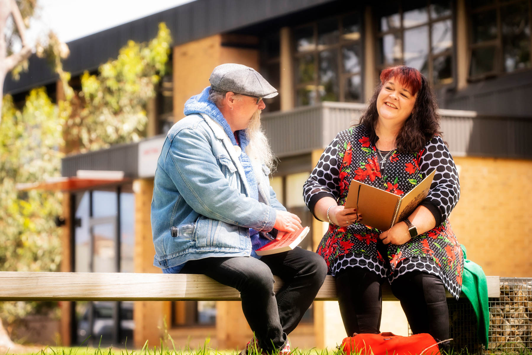 Two students sit outside Uniting College for Leadership and Theology, one is holding a red book, the other an open exercise book