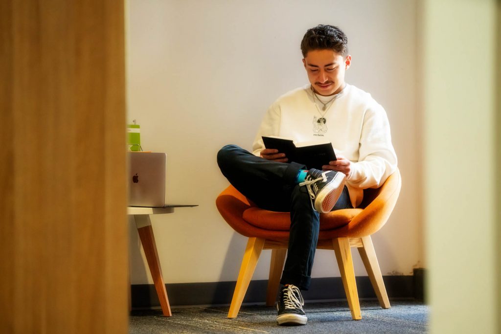 A young Uniting College for Leadership and Theology student in white jumper reads a book, as his laptop lays open on a side-table next to him