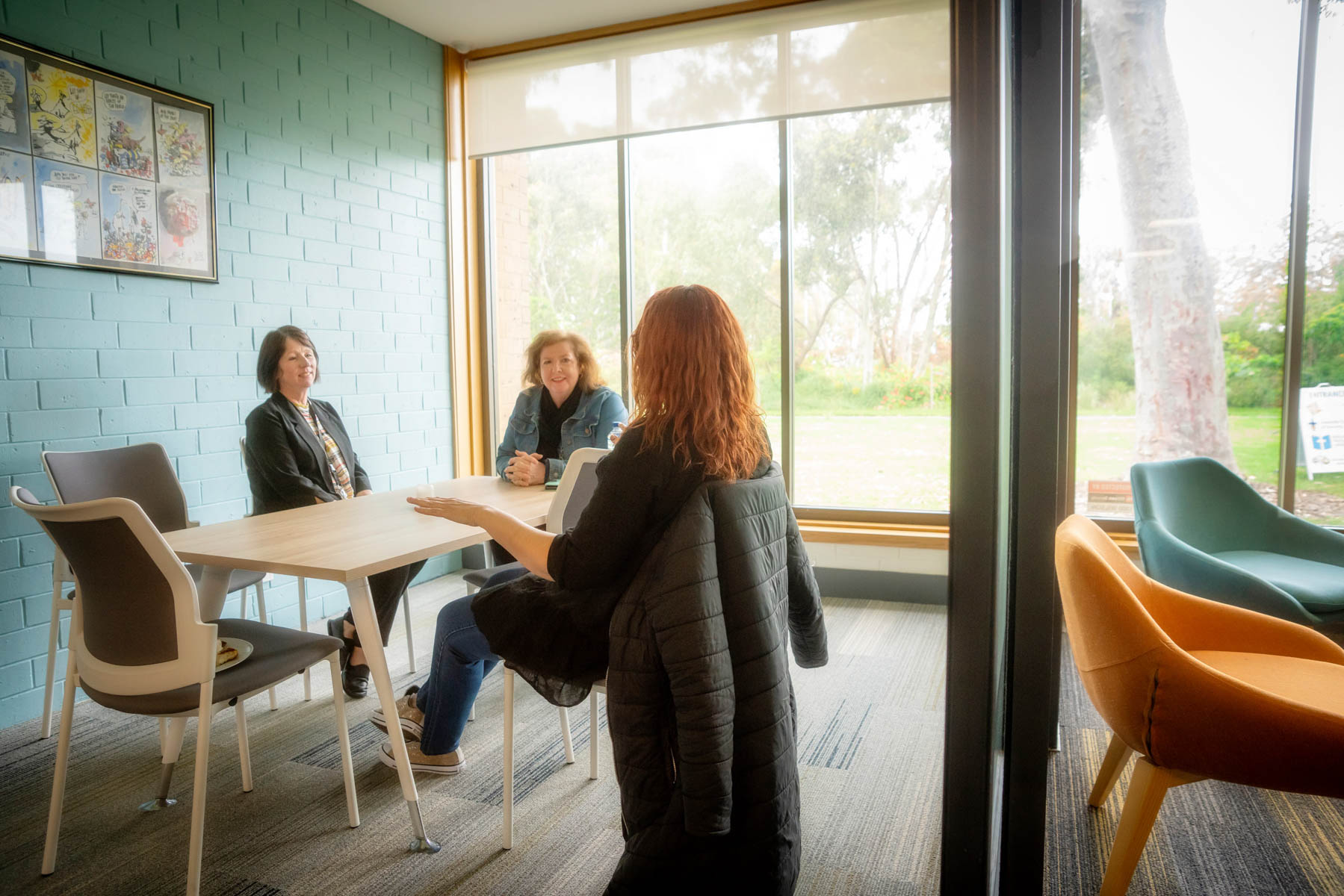 Three Uniting College for Leadership and Theology students sit around a table chatting. There is a large window overlooking the garden in the background.