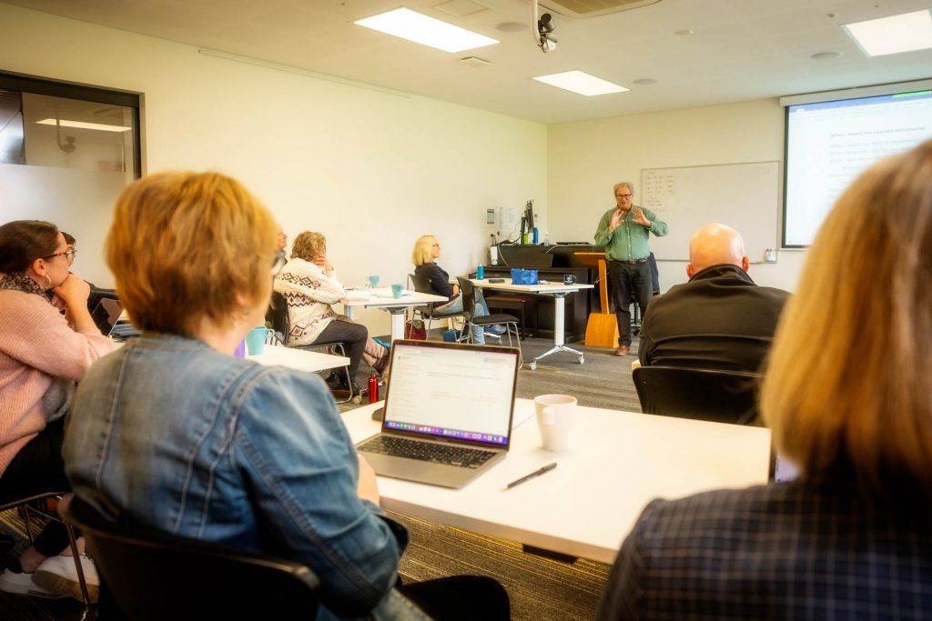 A lecturer in a green shirt holds his hands open to a classroom full of listening students—the student in the foreground has written notes on an open laptop