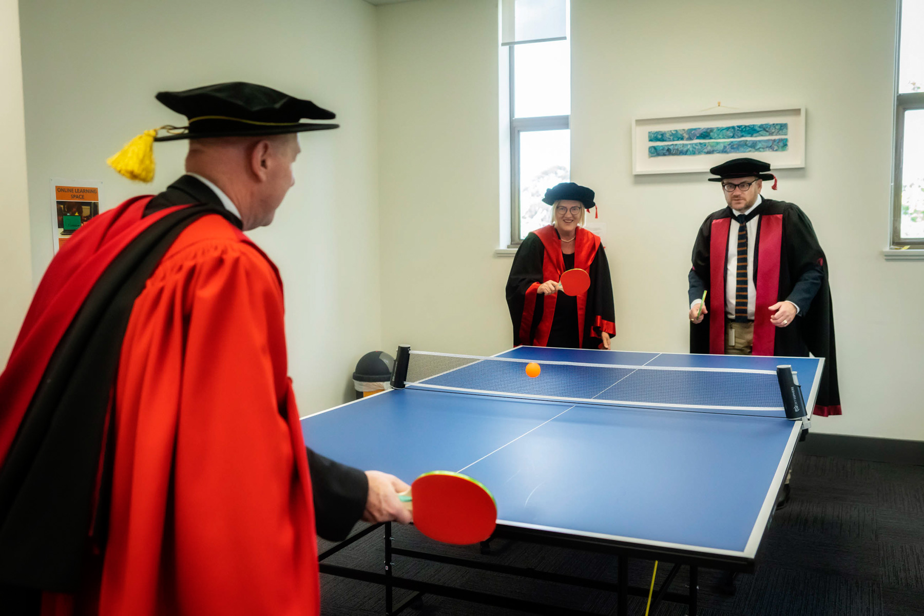 Three postgraduate students graduating from Uniting College for Leadership and Theology play table tennis in academic garb.