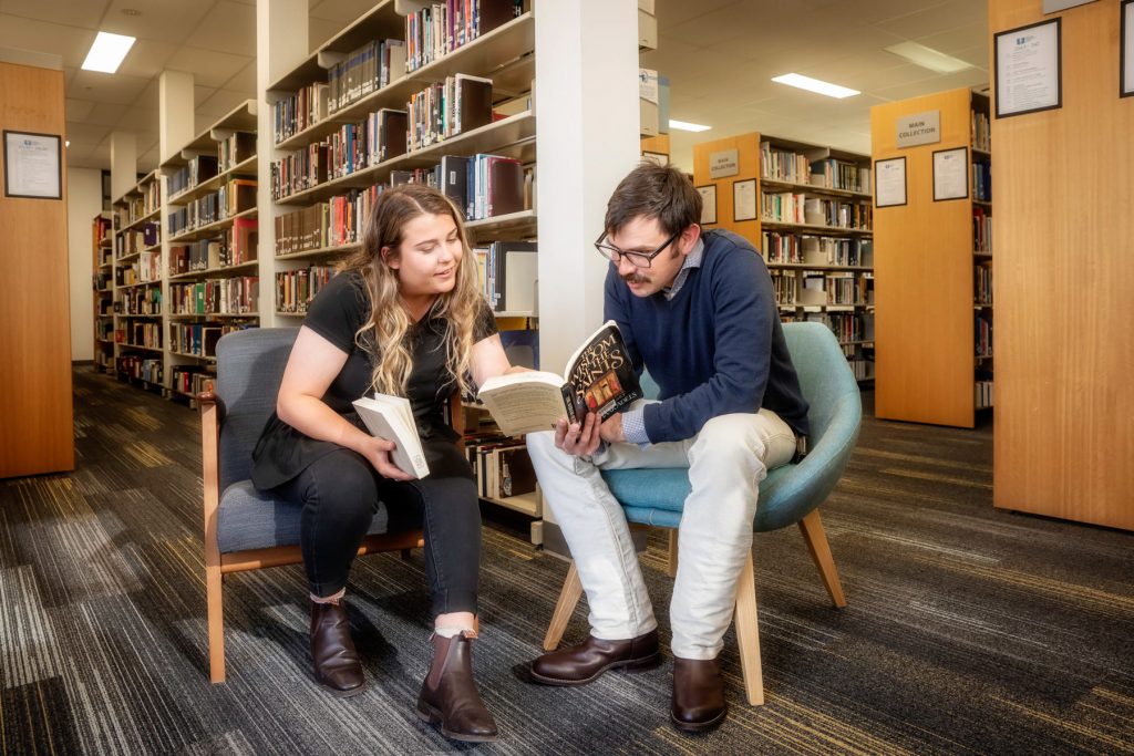 A student from Uniting College for Leadership and Theology points to a page from another student's book, as they sit together on library chairs, amid full book shelves.