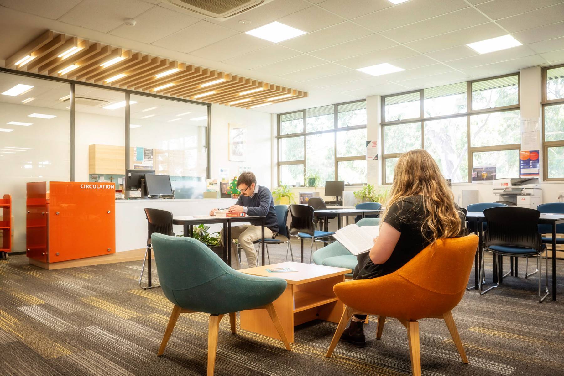 A student in black glasses from Uniting College for Leadership and Theology reads a red book at a library table, as another student reclines on an orange chair