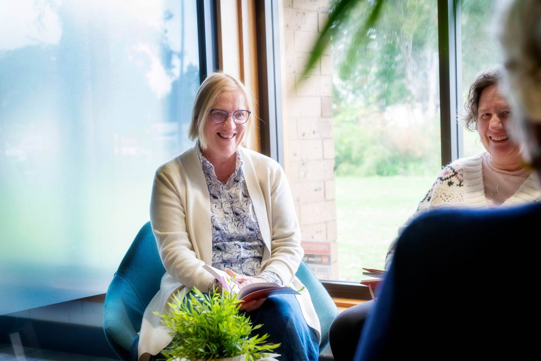 Three Uniting College for Leadership and Theology students sit around a table chatting, one student wearing glasses is holding a book open in her lap.