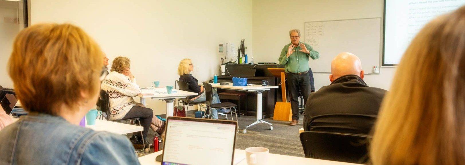 A lecturer in a green shirt holds his hands open to a classroom full of listening students—the student in the foreground has written notes on an open laptop