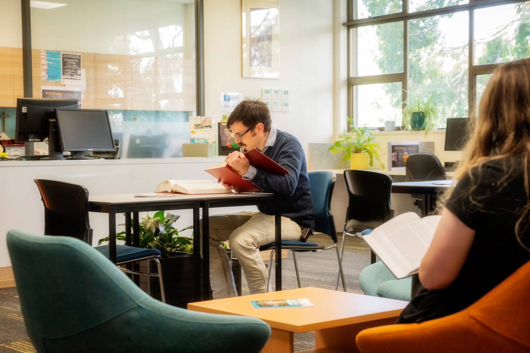 A student in black glasses from Uniting College for Leadership and Theology reads a red book at a library table, as another student reclines on an orange chair